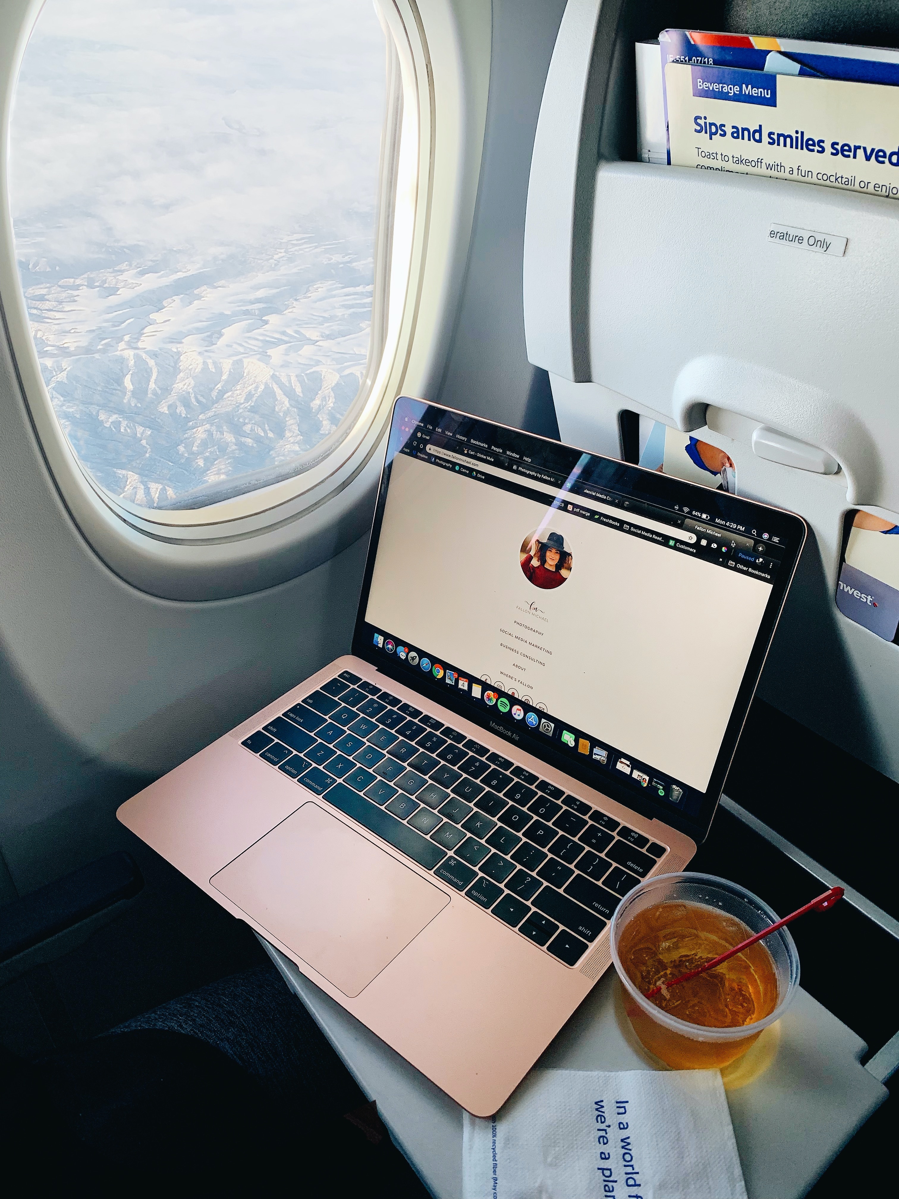 MacBook open on the tray table of an airport seat. There is a drink next to the laptop and the window is opened with a view of mountain peaks. 