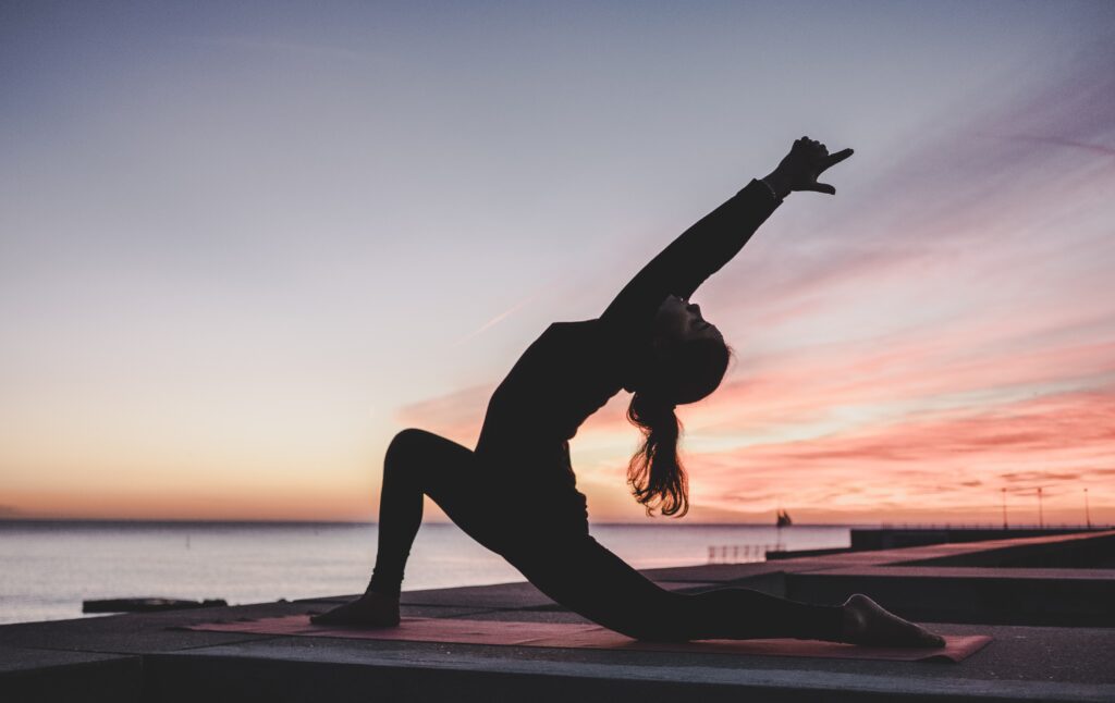 girl doing yoga in the sunset on the beach. The water is in the background and there is a silhouette of the girl stretching.
