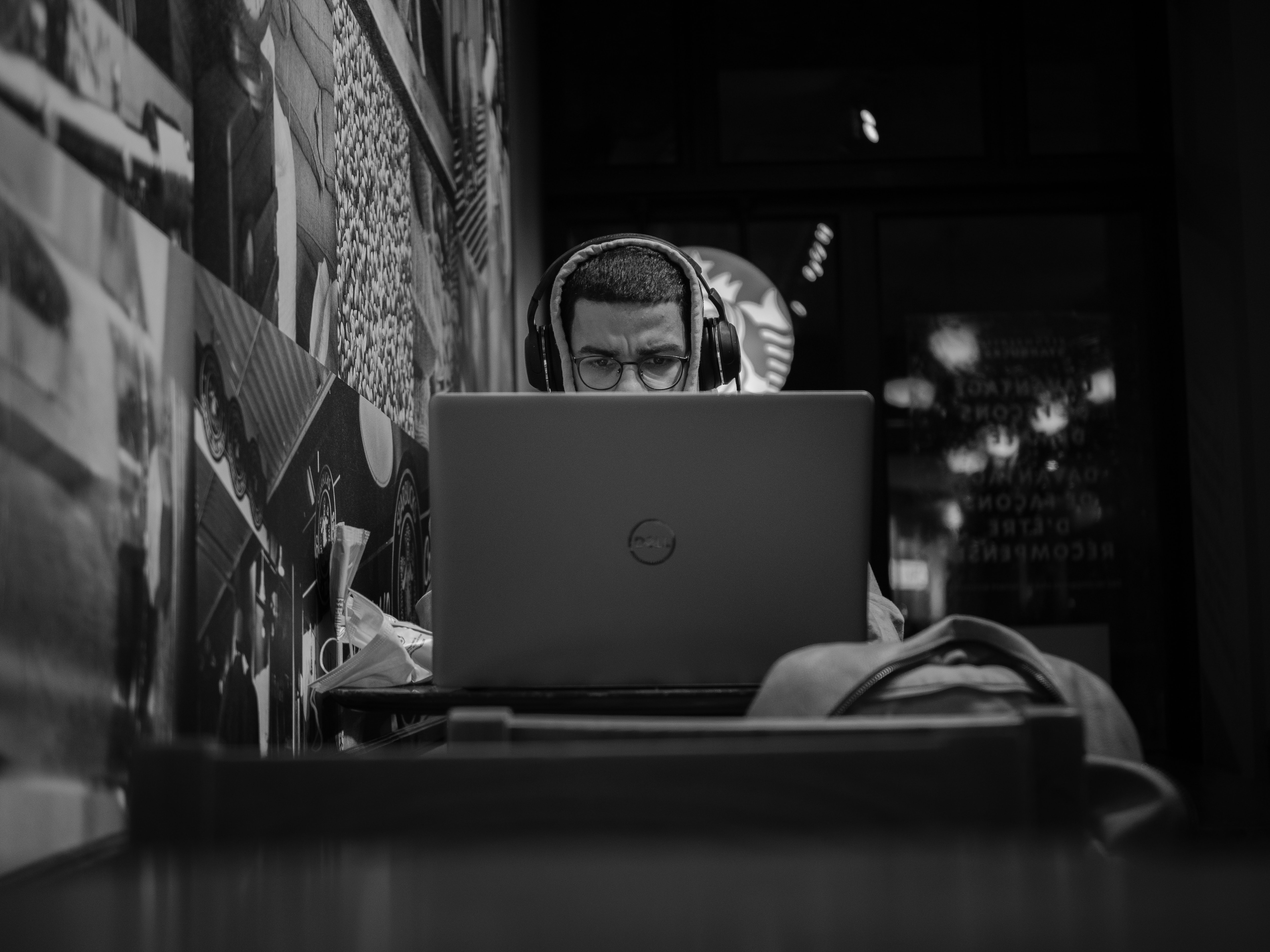 black and white image of a man in a coffee shop working on his laptop. He has headphones on and the computer is covering half of his face,