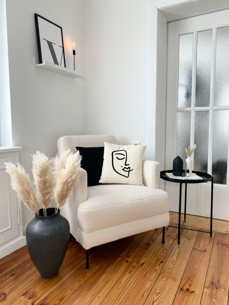 living room corner with white armchair and a black vase next to it. The armchair has two pillows, one black and another with a face on it