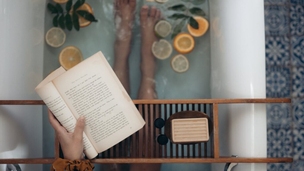 Person Holding Book Near Brown Wooden Table. She is in a bath as a Sunday self-care activity