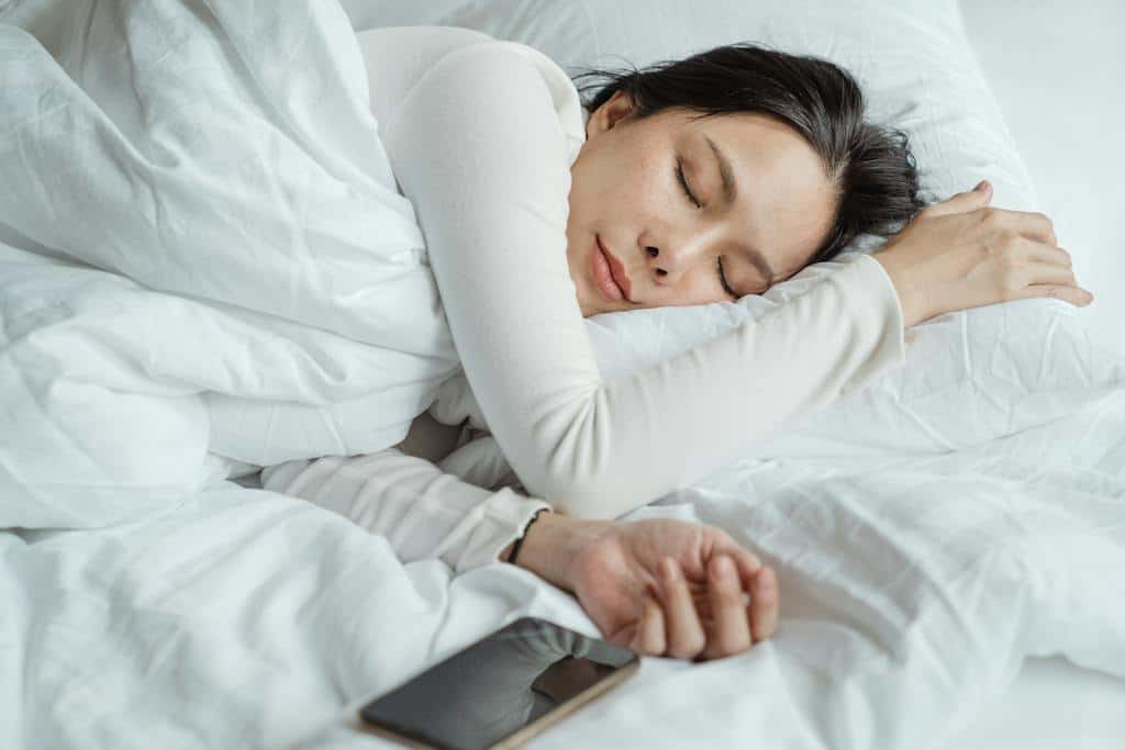 Calm Asian female wearing white pajama sleeping in comfortable bed with white sheets near modern mobile phone in morning. She is getting a good night's sleep.